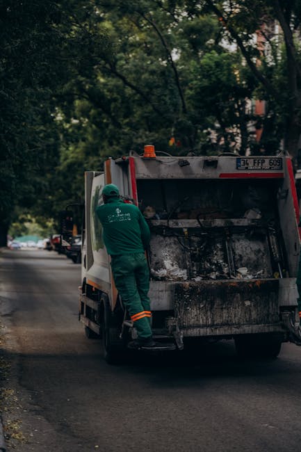 A sanitation worker wearing a green uniform and helmet is standing next to a large, black and white refuse collection vehicle parked on the side of a paved residential street. The vehicle's rear compactor section is open, revealing accumulated waste and debris within. The street is lined with trees that have dense green foliage, which partially shade the scene. Several parked cars are visible further down the road, and the background includes hints of residential buildings behind the trees. The lighting appears natural, indicating daytime with slight overcast conditions, and the scene captures the routine of waste collection services, indicative of an alternative private rubbish removal or on-site clearance activity by a company such as Rubbish Clearance Balham, supporting efficient rubbish disposal without reliance on local authority services.