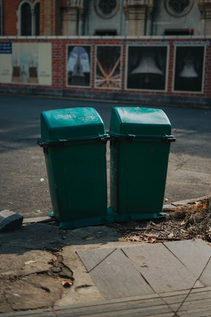 Two green plastic wheelie bins are positioned side by side on a paved sidewalk, with their lids closed and secured with black clips. The bins have smooth, matte surfaces and appear to be made of durable, weather-resistant plastic. They are situated on a curb edge, with some scattered debris and dried leaves nearby. Behind the bins, a residential street is visible, featuring a brick building with decorative window frames and a mural or advertisement painted on a wall, though the details are partially out of focus. The ground in front of the bins is uneven, with a section of cracked pavement and a small section of curb. Natural light illuminates the scene, casting soft shadows and enhancing the muted, neutral tones of the surroundings. The image subtly relates to private waste collection or rubbish removal services, in line with the context of alternative waste handling solutions.