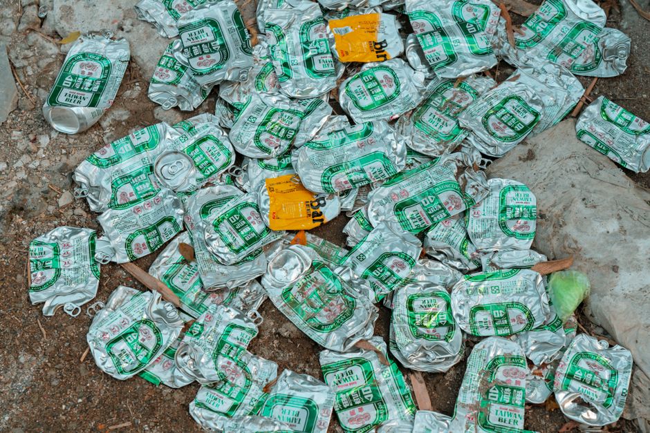 A collection of crushed aluminium cans scattered on the ground, predominantly silver in color with green and white labels indicating Taiwan Beer branding. The cans are crumpled and piled together on a dirt surface mixed with small stones, dried leaves, and scattered pieces of trash. Some cans are partially obscured, with a few showing the pull-tab openings intact. Among the cans, there is a small yellow label and a partially visible plastic bag containing more waste. The environment appears outdoor, possibly a street or alleyway, with ambient natural light highlighting the metallic sheen of the cans and the rough texture of the ground. This scene reflects typical discarded waste that may be handled through private rubbish clearance services, such as those provided by Rubbish Clearance Balham, especially when arranging independent collection or on-site clearance for recyclable materials.