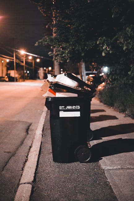 A black wheeled waste bin with a white label and the name 'St. John's' printed on it is positioned on the pavement beside a sidewalk at night. The bin's lid is open and filled with a variety of rubbish, including cardboard boxes and paper waste. The nearby street is illuminated by warm streetlights, casting shadows across the road and the dark, leafy trees lining the pavement. In the background, additional streetlights and parked vehicles are visible, creating a quiet, urban atmosphere. The scene suggests an example of private or alternate waste collection, with the waste bin prepared for collection or disposal, consistent with rubbish removal services such as those offered by Rubbish Clearance Balham. The textured asphalt, the partially visible curb, and the surrounding greenery reinforce the setting in a residential or semi-rural street environment, illustrating the typical urban waste dispersal scene at night.