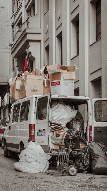 A white panel van parked on a city street with its rear doors open, revealing a large load of mixed rubbish including black trash bags, flattened cardboard boxes, and wooden pallets inside and on top of the vehicle. Several black trash bags and white rubbish sacks are stacked on the ground near the van, with a metal trolley and a small hand cart also visible in front of the open doors. The surrounding environment features a multi-storey building with grey, smooth concrete facade, narrow windows, and architectural detailing typical of urban residential or commercial properties. The scene appears to be in the process of a private waste removal or estate clearance, with the rubbish likely collected for disposal by a professional service such as Rubbish Clearance Balham, as indicated by the context of the website. The lighting suggests daytime with soft, natural light illuminating the scene, emphasizing the textures of the rubbish bags, cardboard, and building exterior. The overall setup underscores the nature of independent collection efforts in urban areas, aligning with services that offer alternative waste handling and rubbish removal outside of local authority collections.