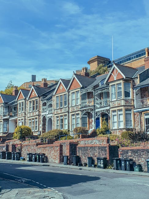 A row of Victorian-style terraced houses with ornate white metalwork balconies, bay windows, and pitched roofs, situated along a paved street with a brick boundary wall and a line of black waste bins at the curb. The buildings are constructed from red brick with contrasting light-colored trims, and small landscaped front gardens with shrubs and greenery are visible in front of each property. The sky is clear and blue, suggesting a bright, sunny day. The scene reflects a typical residential area where private waste collection or rubbish removal services might be employed, supported by the presence of waste bins alongside the curb, indicating ongoing rubbish clearance or disposal activities perhaps coordinated by local or independent waste management providers like Rubbish Clearance Balham.