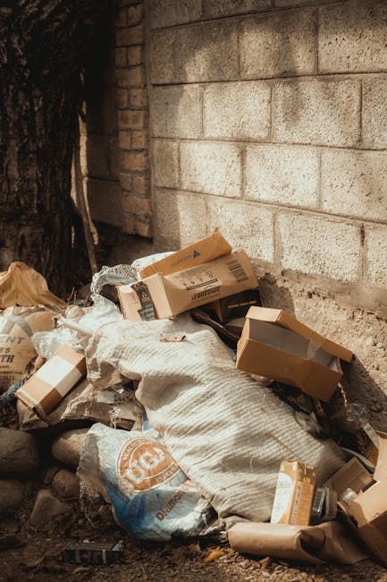 A pile of mixed waste materials is situated at the base of a rough, beige concrete block wall, partially shaded by a nearby tree with dark bark. The debris includes flattened cardboard boxes, some with visible printed labels, and various plastic bags, one of which appears to be a branded shopping bag, all in neutral tones such as brown, white, and light tan. The cardboard boxes are crumpled and some are torn, with edges folded or leaning against each other in a disorderly heap. The large white sack, made of woven fabric, lies among the smaller refuse, with a textured surface that contrasts with the smooth, glossy finish of the boxes. The ground beneath the rubbish consists of small rocks and dirt, indicating an outdoor setting near a building or land clearance area. The scene exemplifies typical waste accumulation that could be associated with privately arranged rubbish removal services like those offered by Rubbish Clearance Balham, highlighting the kind of waste handled during on-site clearance or alternative disposal methods away from municipal collection.
