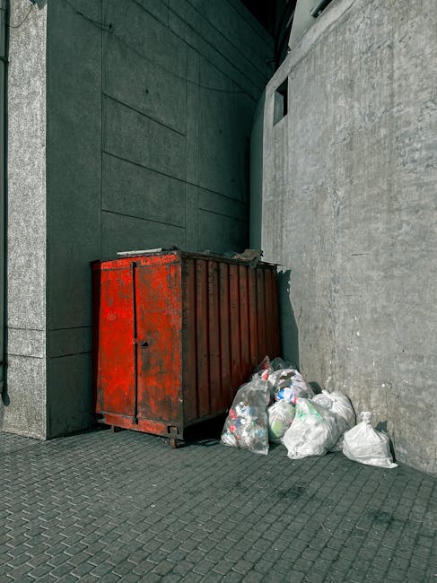 A large, weathered red metal dumpster with visible rust and chipped paint, positioned at the corner of a paved outdoor area between a dark green wall with horizontal paneling and a light gray concrete wall. In front of the dumpster, several white plastic rubbish bags, some slightly crumpled and bulging, contain mixed waste materials. The brownish-black cobblestone paving extends beneath and around the objects, with subtle dirt and debris visible. The scene is illuminated by natural daylight, casting soft shadows and highlighting the textured surfaces of the walls, dumpster, and bags. This setting reflects a typical external rubbish storage area that might be temporarily used for private waste disposal or alternative rubbish collection, as part of a service without involving local authority collection. Rubbish Clearance Balham occasionally handles similar on-site waste removal tasks, ensuring proper clearance of household or commercial waste outside of regular bin collection days.